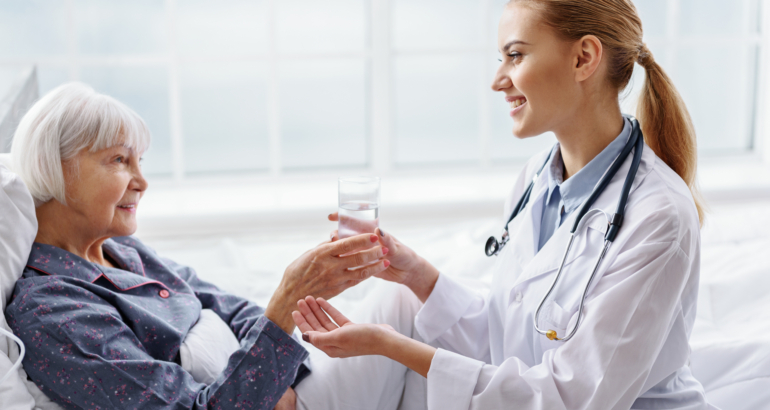 Outgoing young physician holding out to drink old woman while sitting near her on bed in hospital apartment