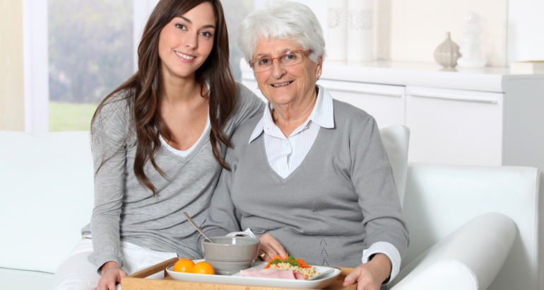 Elderly woman and home carer sitting in sofa with lunch tray